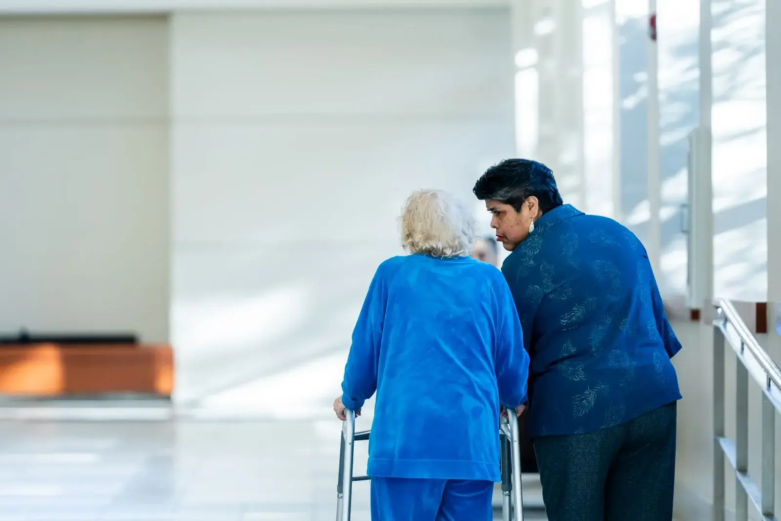 A clinician walking beside an older woman using a walker in a bright clinic corridor